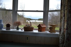 A window with a view of a lake and trees, a wooden shelf with a basket, a birdhouse, and a vase. - Quiet and Cozy Seaside Cottage with Amazing Sunsets (Steuben)