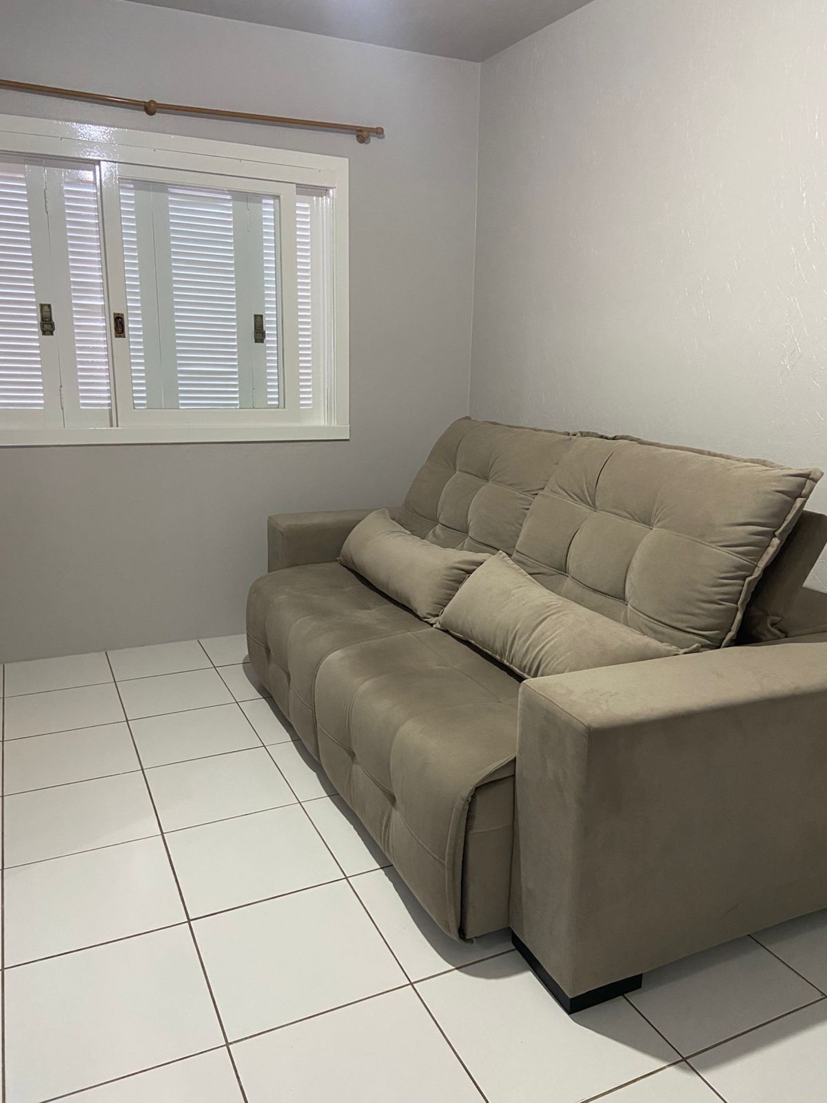 Ein beiges Sofa in einem Raum mit weiß gefliestem Boden und einem Fenster mit heruntergelassenen Jalousien.