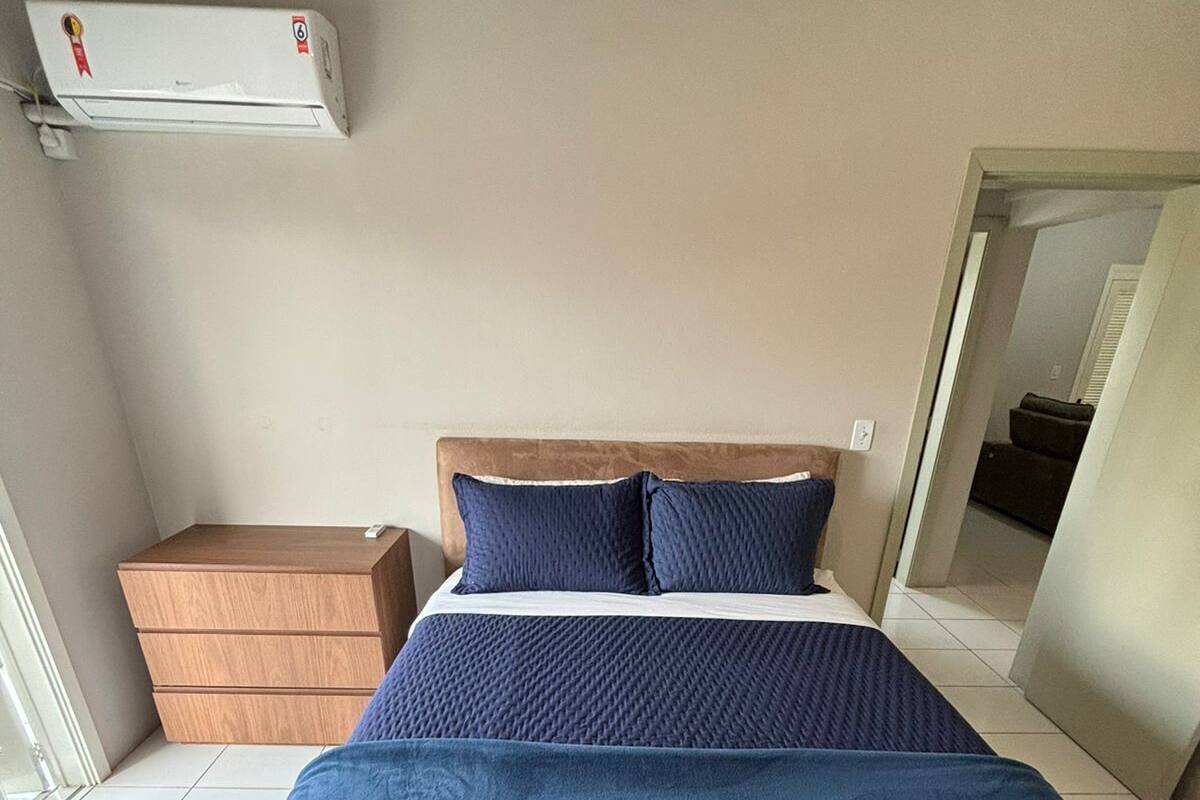 Un dormitorio con una cama grande, una mesita de noche de madera, un equipo de aire acondicionado blanco y vistas a otra habitación a través de una puerta abierta.