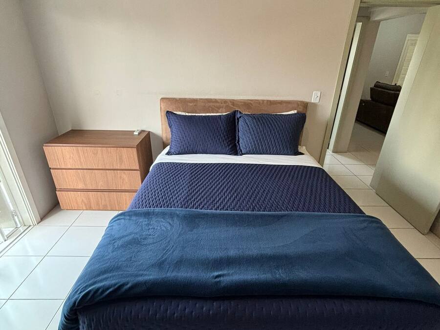 Un dormitorio con una cama grande, una mesita de noche de madera, un equipo de aire acondicionado blanco y vistas a otra habitación a través de una puerta abierta.
