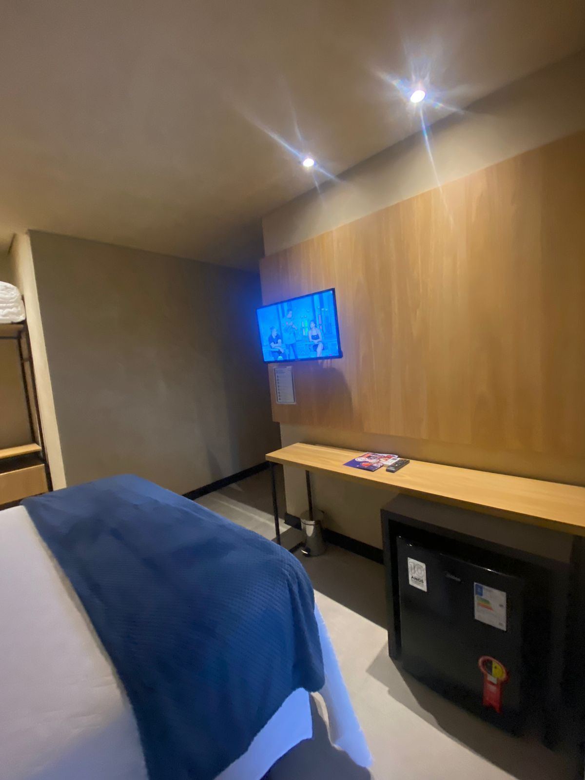Habitació d'hotel amb llit, escriptori, televisió penjada a la paret i un petit frigorífic.