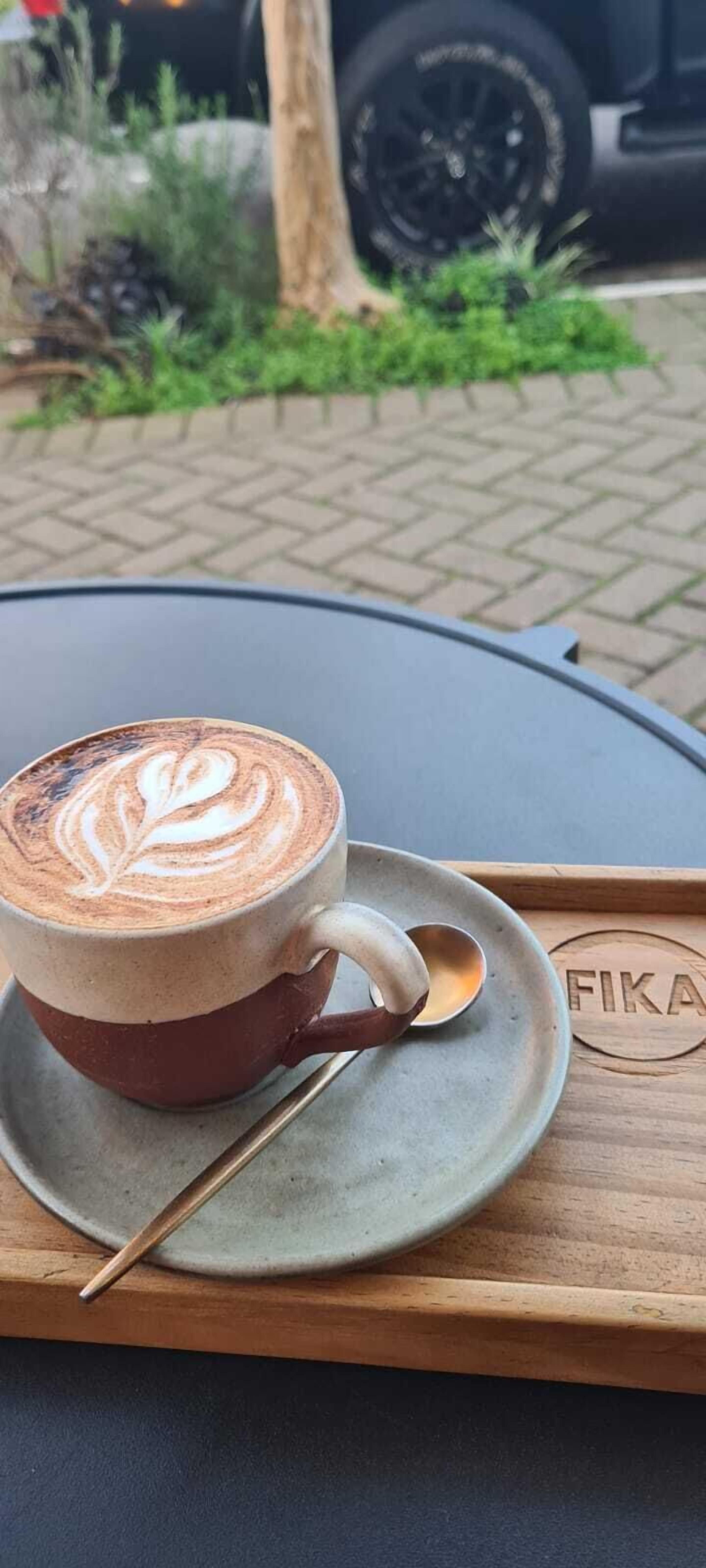 Eine Kaffeetasse mit Latte-Art auf einem Untertasse, platziert auf einem Tablett mit einem Logo, auf einem Tisch im Freien.