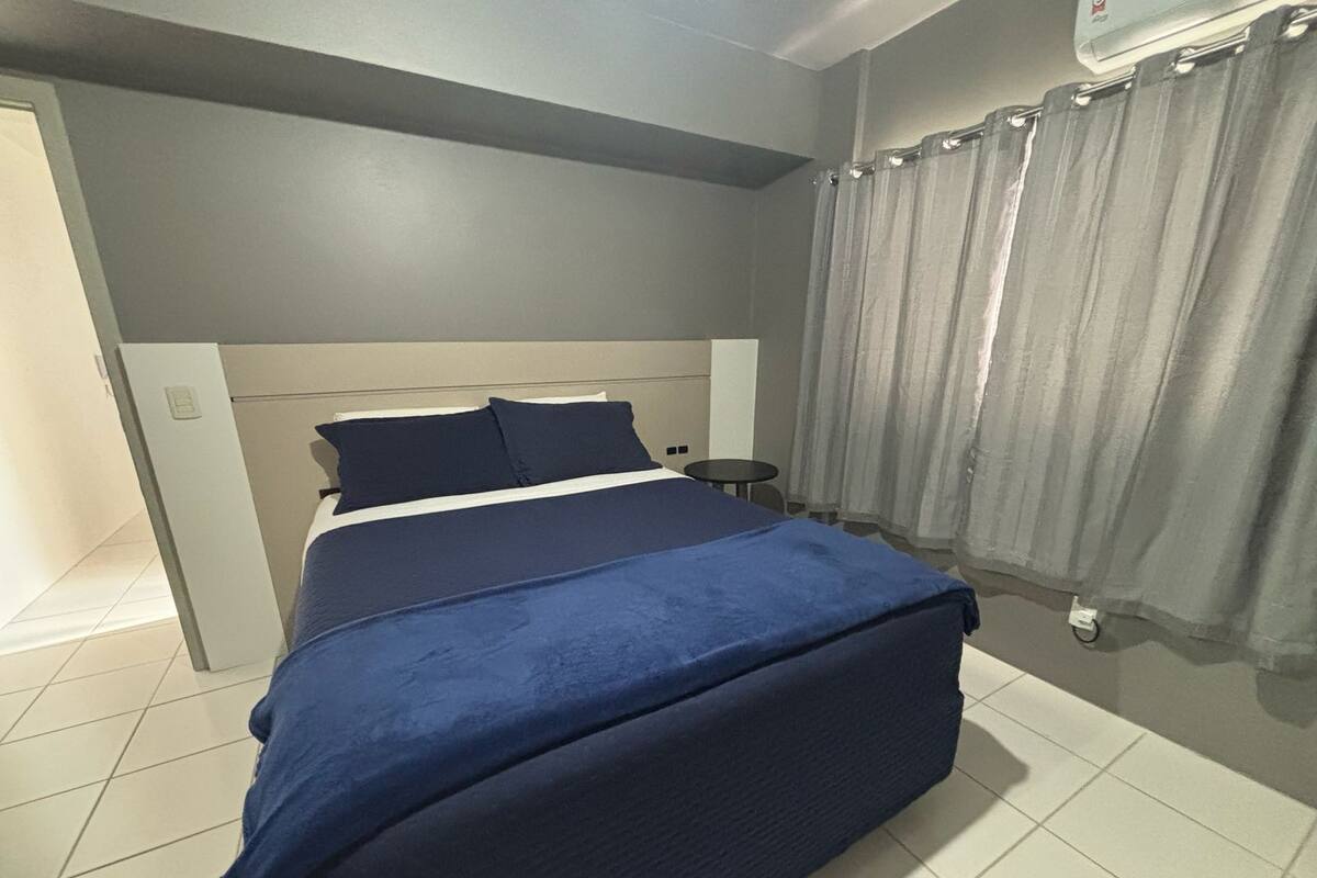 Un dormitorio con cama, mesita de noche, silla y ventana con cortinas.