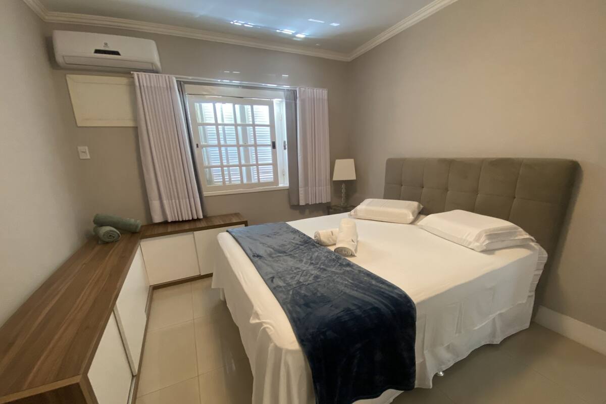 Un dormitorio ordenado con una cama, una mesita de noche, una lámpara y un ventanal con cortinas.