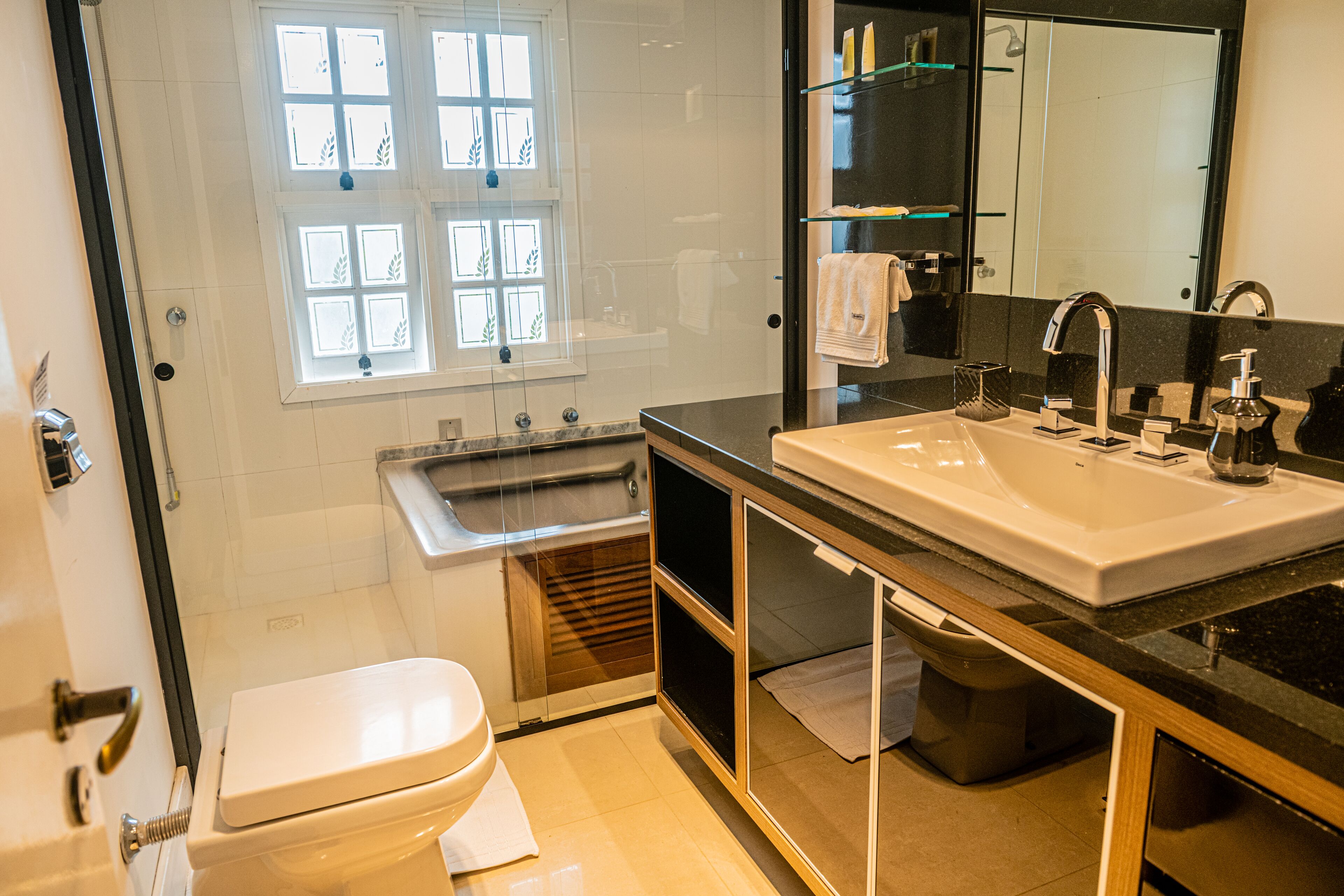 A modern bathroom with a white toilet, a glass-enclosed shower, a large sink with a mirror, and a window with decorative glass panes.