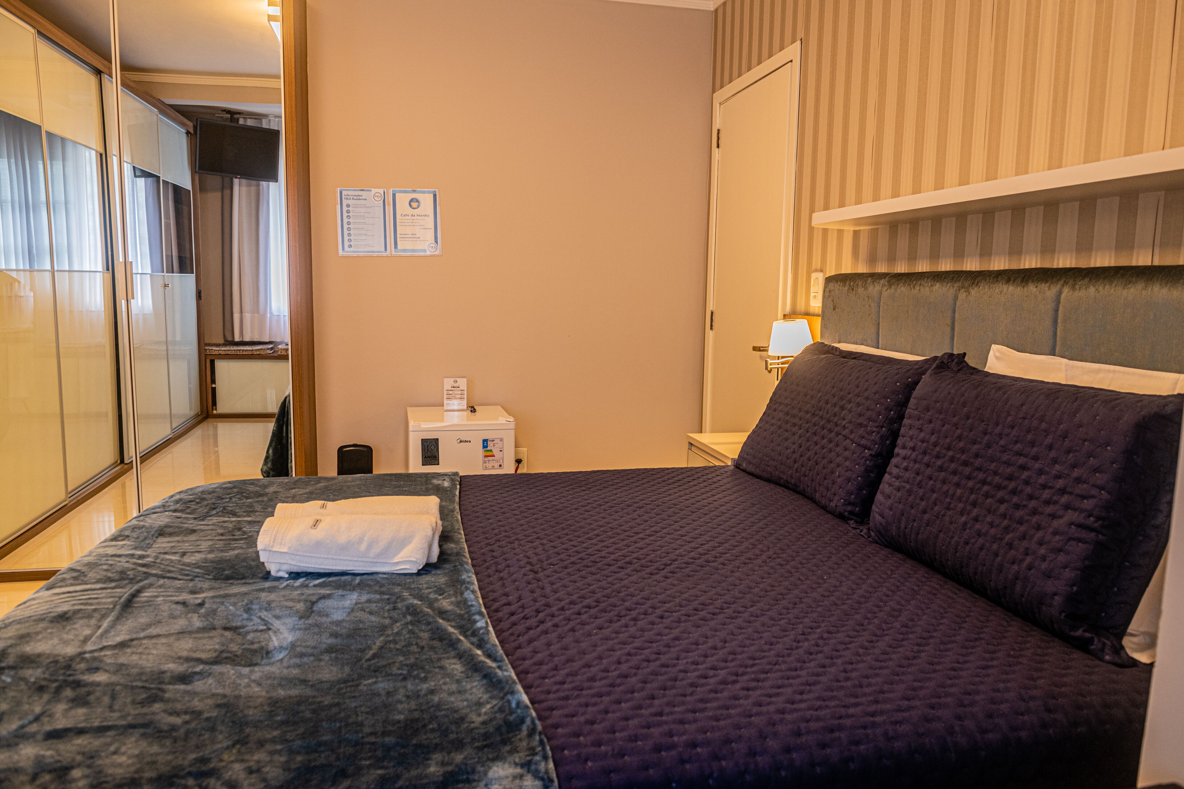 Quarto de hotel com cama, escrivaninha, cadeira, guarda-roupa e mesa de cabeceira.