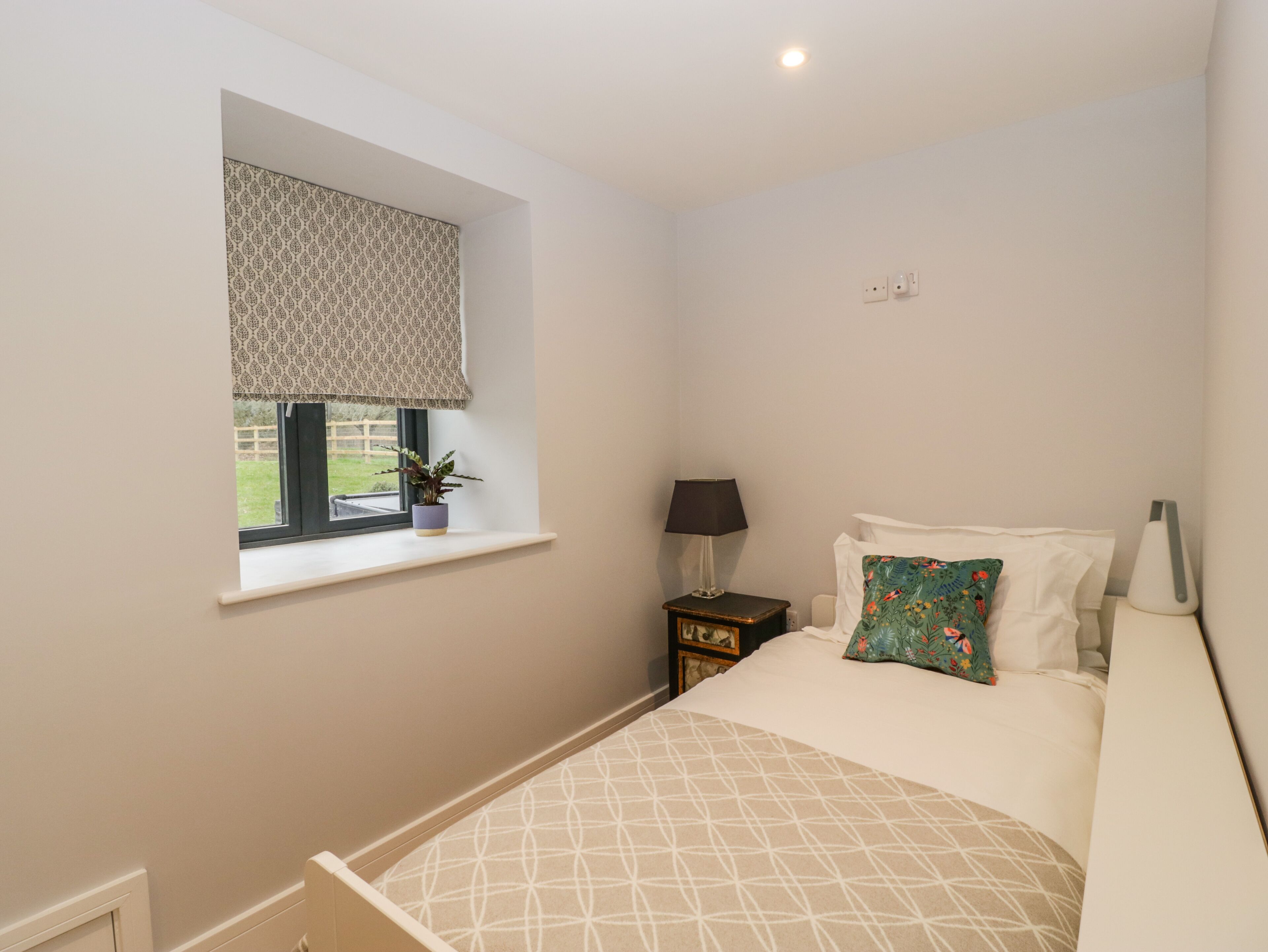 A bedroom with a bed, a window with a patterned blind, a bedside table with a lamp, and a small potted plant.