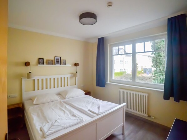 A bedroom with a white bed, a window with blue curtains, and a radiator. - 2-bedroom apartment with a balcony (Ostseebad Binz)