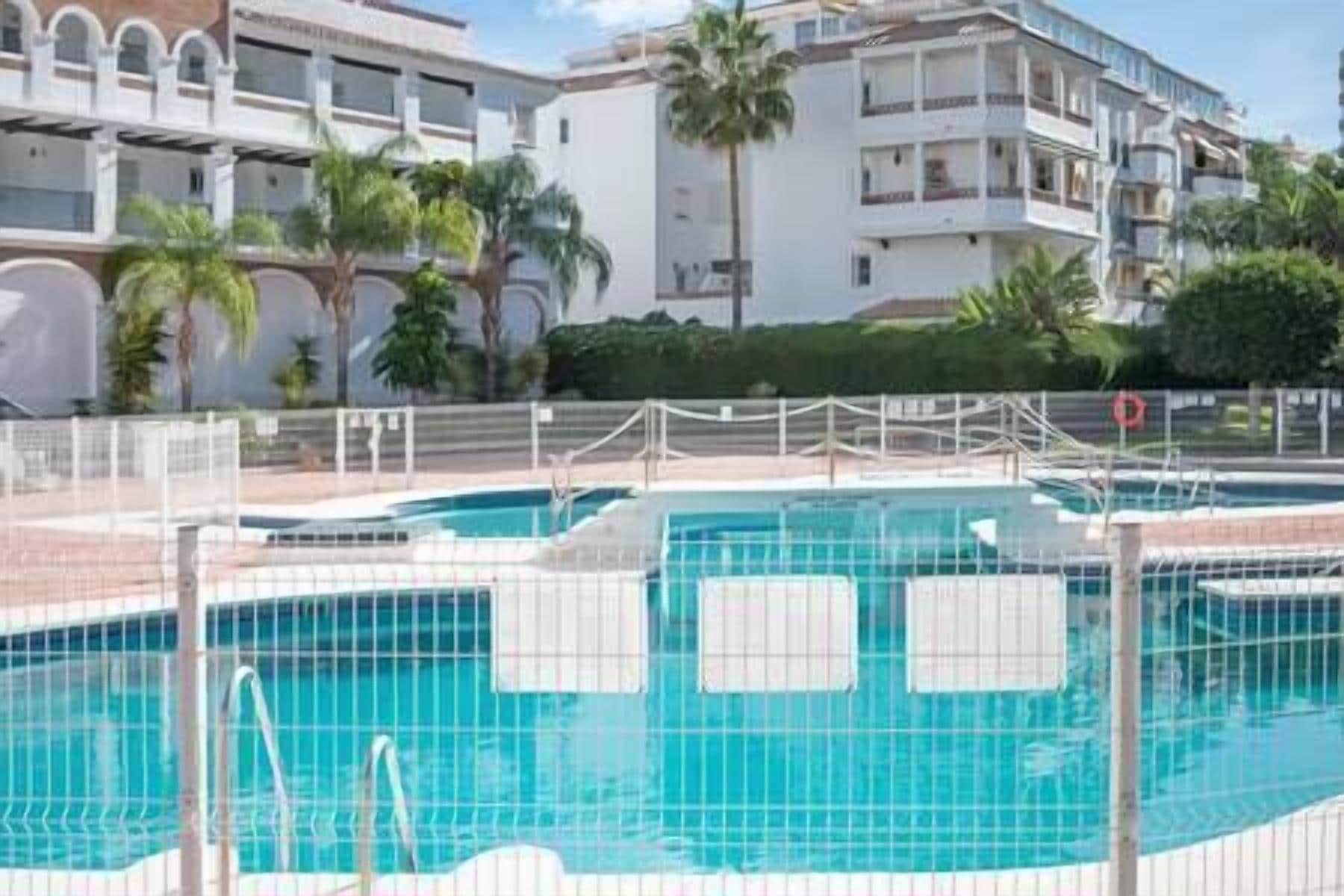 A swimming pool area with multiple pools, surrounded by a fence, and a building with balconies in the background.