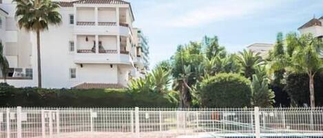 Piscina con cerca blanca, rodeada de palmeras y un edificio con balcones.