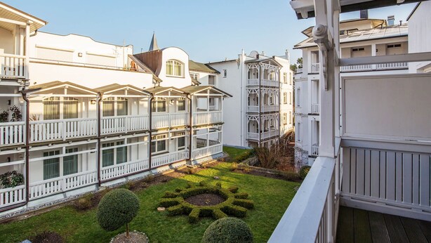 Une cour bordée de bâtiments blancs, un petit jardin et un balcon agrémenté d’une balustrade.
