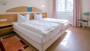 Una camera d'albergo con due letti, una testiera in legno, una finestra con tende, una sedia e un tavolino.