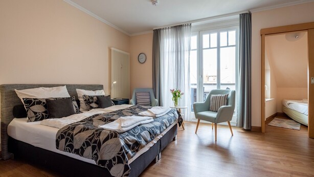 Ein modernes Schlafzimmer mit einem großen Bett, einem Sessel, einem kleinen Tisch mit einer Vase und einem Fenster mit Vorhängen.