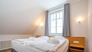 Un lit bien fait dans une pièce dont le plafond est en pente, avec une fenêtre ornée de rideaux, une table de chevet et un luminaire fixé au mur.