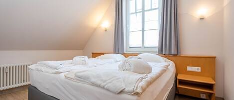 Ein ordentlich bezogenes Bett in einem Zimmer mit schrägverlaufender Decke, einem Fenster mit Vorhängen, einem Nachttisch und einer wandmontierten Lampe.