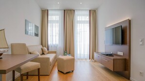Ett modernt hotellrum med trägolv, en soffa, ett matbord, en TV och ett stort fönster med gardiner.