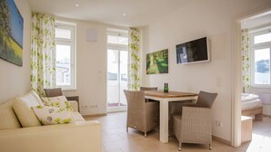 Ein modernes Wohnzimmer mit einer Couch, einem Esstisch und Korbstühlen. An der Wand befinden sich zwei Gemälde, über dem Tisch ist ein Fernseher montiert und ein Fenster mit Vorhängen.