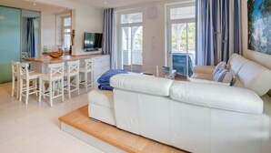 A modern living room with a white leather sofa, a dining area with white chairs, and a kitchen with a TV. - Apartment with sea view, 2 bedrooms and balcony (Ostseebad Binz)