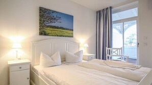 A bedroom with a bed, two bedside tables, a nightstand, a chair, and a large window with curtains. - Apartment with sea view, 2 bedrooms and balcony (Ostseebad Binz)