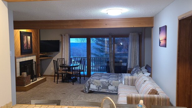 A cozy room with a fireplace, a sofa, a dining table with chairs, and a balcony with a view of trees.