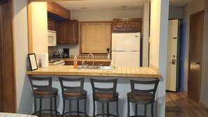 A compact kitchen with wooden cabinets, a built-in microwave, a refrigerator, and a bar area with stools.