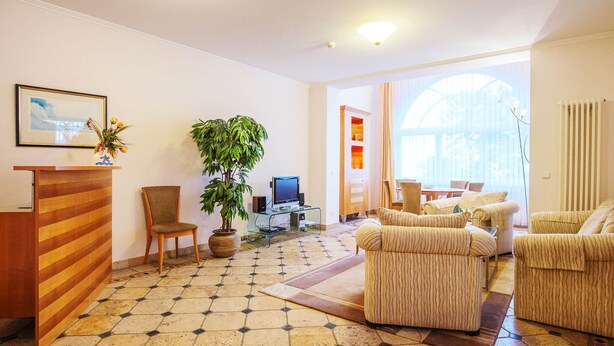 Eine Hotelhalle mit einer Rezeption, einer großen Pflanze im Topf, einem Fernseher und einer Sitzecke mit Sofas.