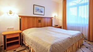Ein Hotelzimmer mit einem großen Bett, einem Holzkopfbrett, zwei Nachttischen, einem Beistelltisch, einem gerahmten Bild an der Wand und einem Fenster mit Vorhängen.