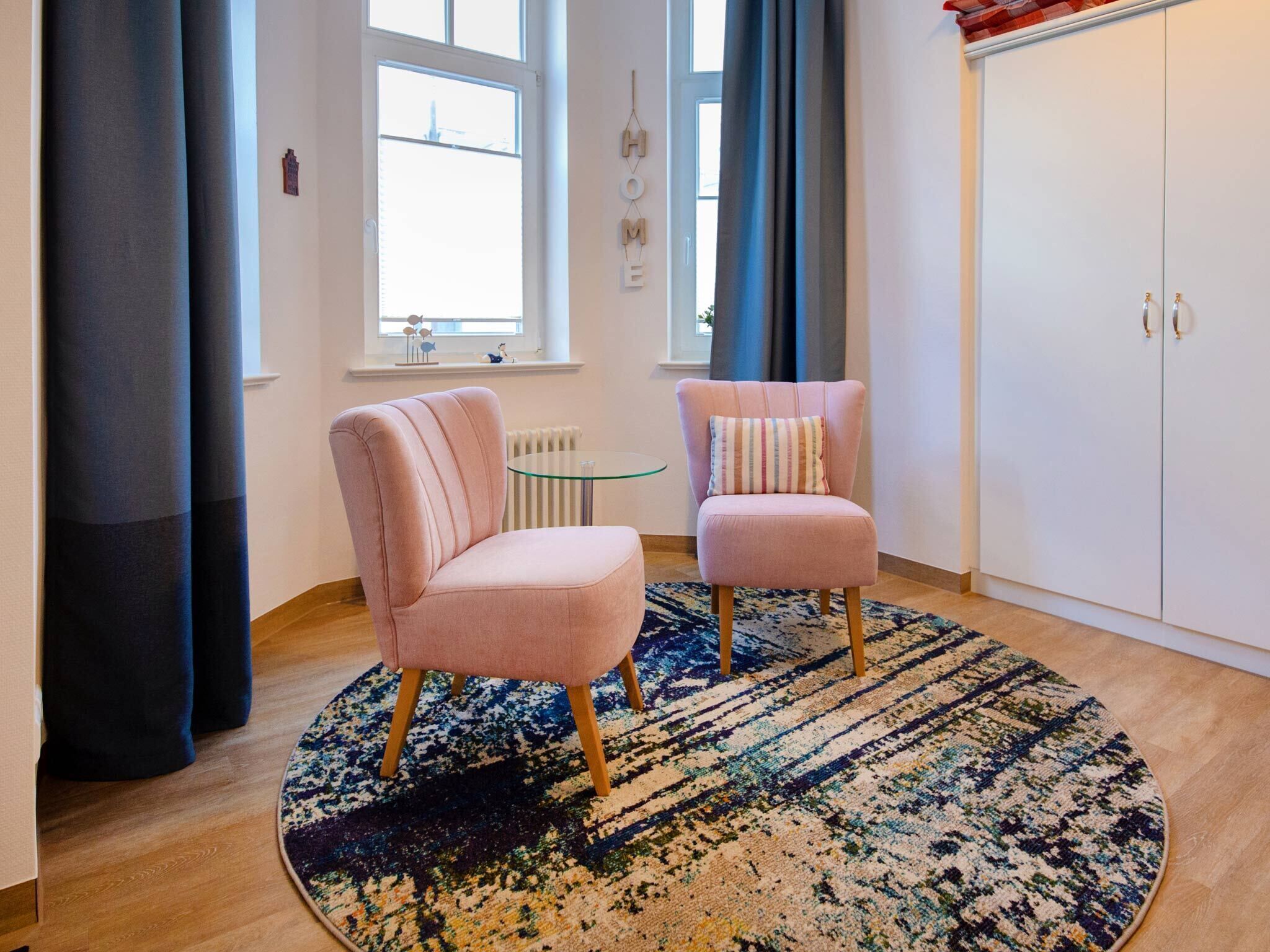 A room with two pink chairs, a glass table, a patterned rug, a white wardrobe, and a window with curtains.