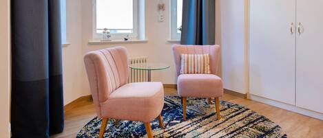 A room with two pink chairs, a glass table, a patterned rug, a white wardrobe, and a window with curtains.