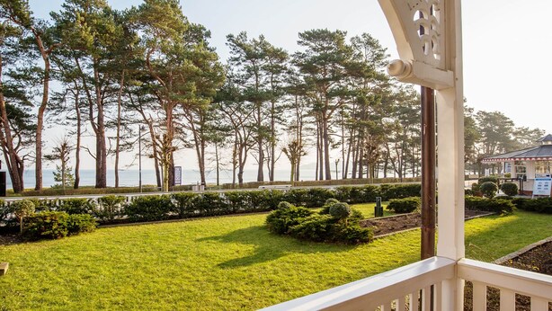 A view from a balcony overlooking a well-maintained lawn with neatly trimmed bushes and tall trees. - Apartment with sea view, 1 bedroom and balcony (Ostseebad Binz)
