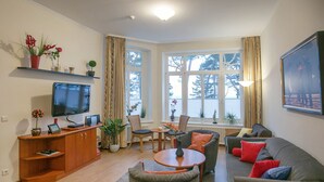 A living room with a TV, seating, a dining table, and a large window with curtains. - Apartment with sea view and 2 bedrooms (Ostseebad Binz)