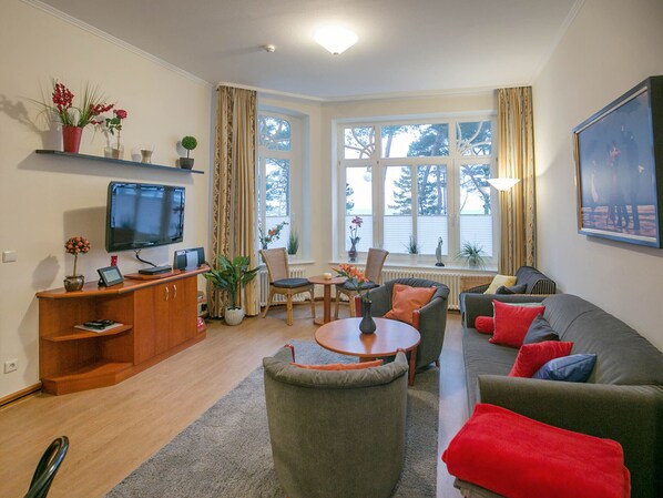 A living room with a TV, seating, a dining table, and a large window with curtains. - Apartment with sea view and 2 bedrooms (Ostseebad Binz)