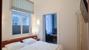 Une chambre d’hôtel avec un lit, une fenêtre avec des stores, une table de chevet, une lampe et un radiateur.