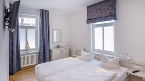 A bedroom with a large bed, a bedside table, a nightstand, a window with blinds, a radiator, and a curtain. - Apartment with sea view and 1 bedroom (Ostseebad Binz)
