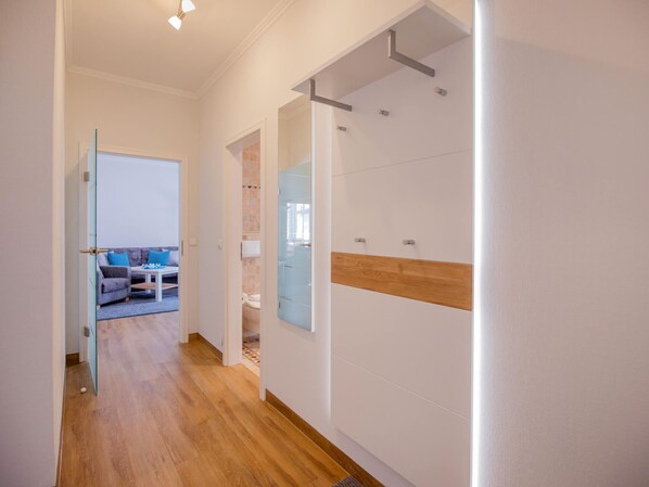 Een gang met een houten vloer, een glazen deur die leidt naar een woonruimte met een bank en een salontafel, en een badkamer met douche en toilet.