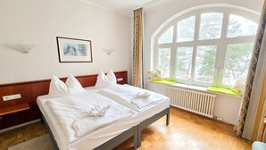 A neatly made bed with pillows and a towel, a framed picture on the wall, a window with a view of trees, and a radiator under the window. - Apartment with sea view and 1 bedroom (Ostseebad Binz)