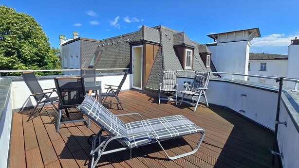 En træterrasse med liggestole og et bord, med udsigt over et boligområde med et hus og et træ.