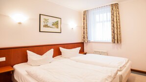 Hotellihuone, jossa on kaksi sänkyä, puinen pääty, seinällä kehystetty taulu ja ikkuna, jossa on verhot.