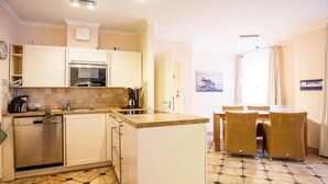 Eine kompakte Küche mit Geschirrspüler, Backofen und Kaffeemaschine. Es gibt eine Essecke mit Korbstühlen und einem Tisch. Der Boden weist ein strukturiertes Fliesenmuster auf.