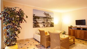 Ein Wohnzimmer mit einer Couch, zwei Korbstühlen, einem Couchtisch, einer großen Topfpflanze, einem Bild mit einer Strandansicht und einem Fernseher auf einer Holzkommode.