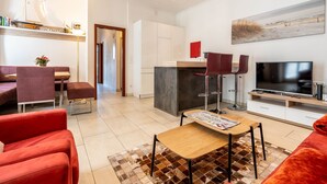Una sala de estar moderna con televisor, zona de estar y cocina al fondo.