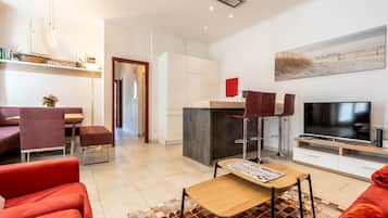 A modern living room with a TV, seating area, and a kitchen in the background.