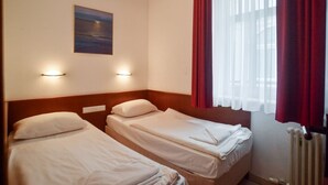 Habitación de hotel con dos camas individuales, una mesita de noche, una lámpara de pared, un cuadro y una ventana con cortinas rojas.