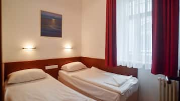A hotel room with two single beds, a nightstand, a wall-mounted lamp, a painting, and a window with red curtains.