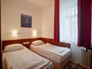 Ein Hotelzimmer mit zwei Einzelbetten, einem Nachttisch, einer Wandlampe, einem Gemälde und einem Fenster mit roten Vorhängen.