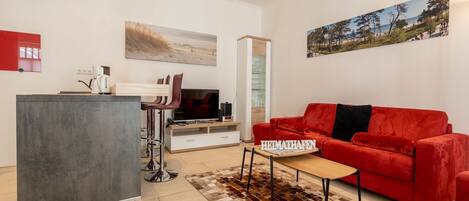 Ein modernes Wohnzimmer mit einem roten Sofa, einem Couchtisch und einem Fernseher. Es gibt eine Küchenzeile mit einer Bar und einem roten Schrank.