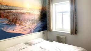 Une chambre avec une grande fresque murale représentant un coucher de soleil sur une plage, un lit avec du linge de maison blanc et une fenêtre avec des rideaux.