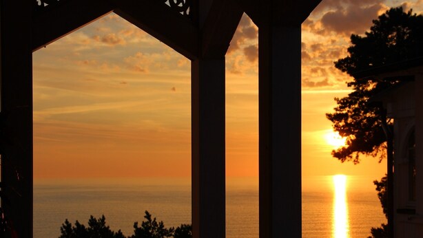 A sunset view through a window with a view of the sea and trees. - Apartment with sea view, 2 bedrooms and balcony (Ostseebad Binz)