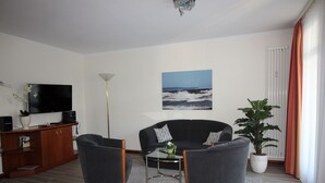 A living room with a sofa, two armchairs, a coffee table, a TV, a floor lamp, a potted plant, and a painting of the sea. - Apartment with sea view, 2 bedrooms and balcony (Ostseebad Binz)