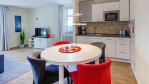 Une chambre d’hôtel moderne dotée d’un coin repas avec une table ronde et des chaises rouges, d’une kitchenette équipée d’un four à micro-ondes et d’un évier, et d’un coin salon avec un téléviseur et une plante.